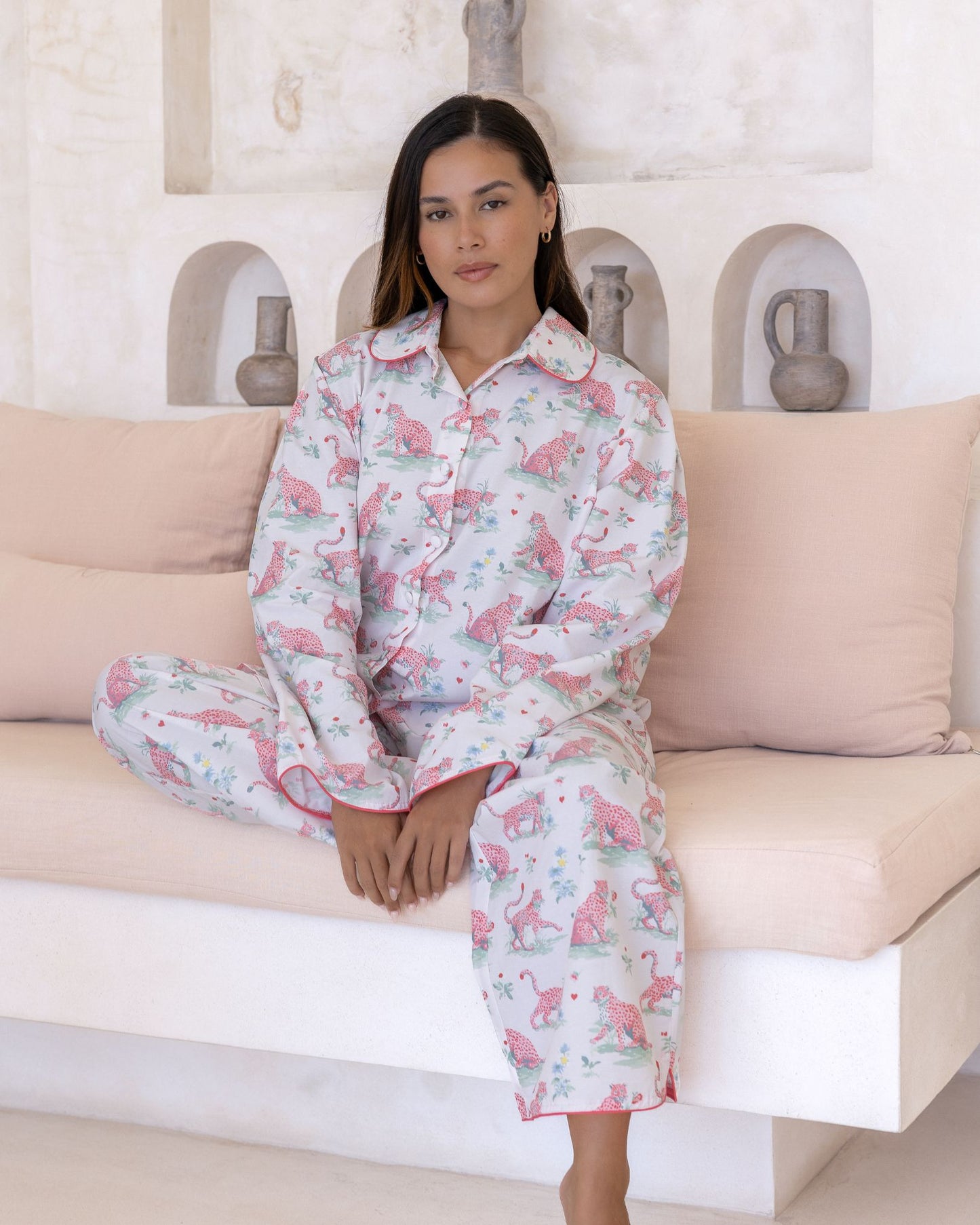 Layla Pink Long PJ Set – model seated on sofa with hands resting in lap, wearing white long-sleeve pyjama set with pink cheetah print and pink piping detail